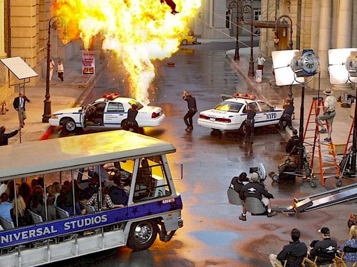 A film set with an explosion, police cars, stunt performers, filming equipment, and a Universal Studios tour tram carrying spectators.