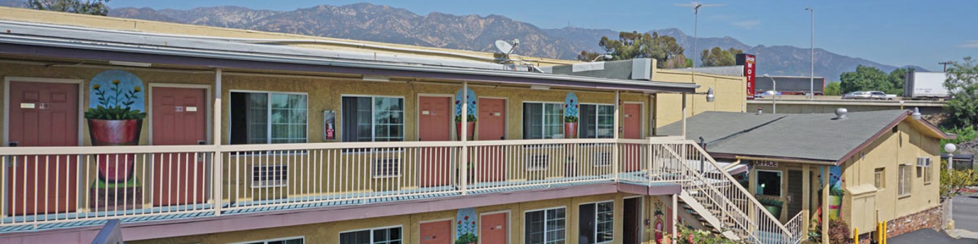 The image shows a two-story motel with exterior corridors, colorful doors, and outdoor staircases. There are mountains in the background and parked cars visible.