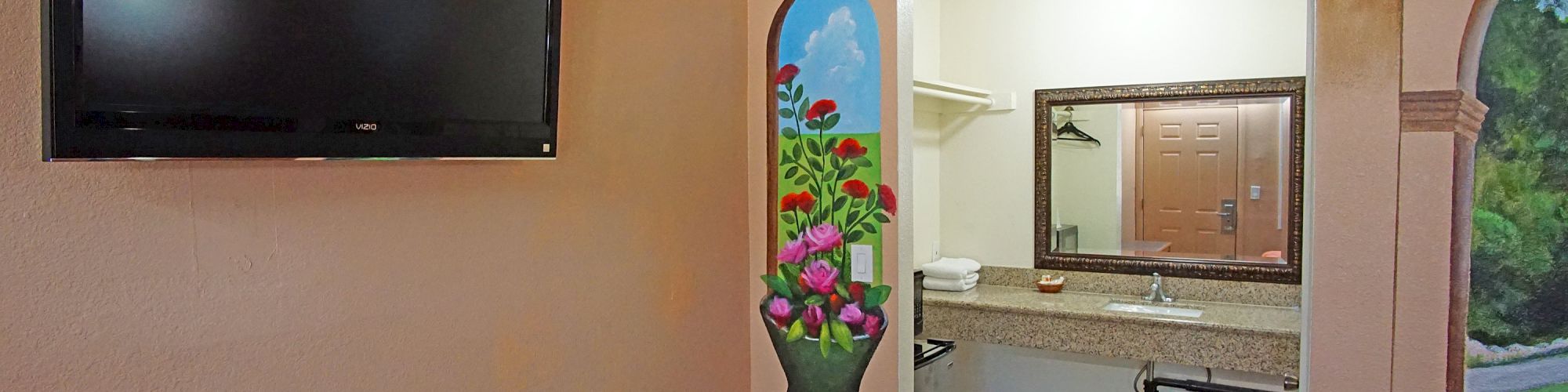 The image shows a hotel room with a wall-mounted TV, a mural of a vase with flowers, a vanity area with a large mirror, and a bathroom door.