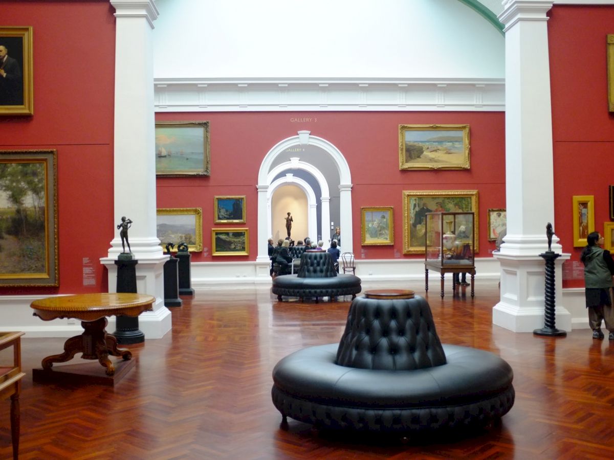 The image shows an art gallery with red walls, various paintings, sculptures, and black round seating in the center of a room with a wooden floor.