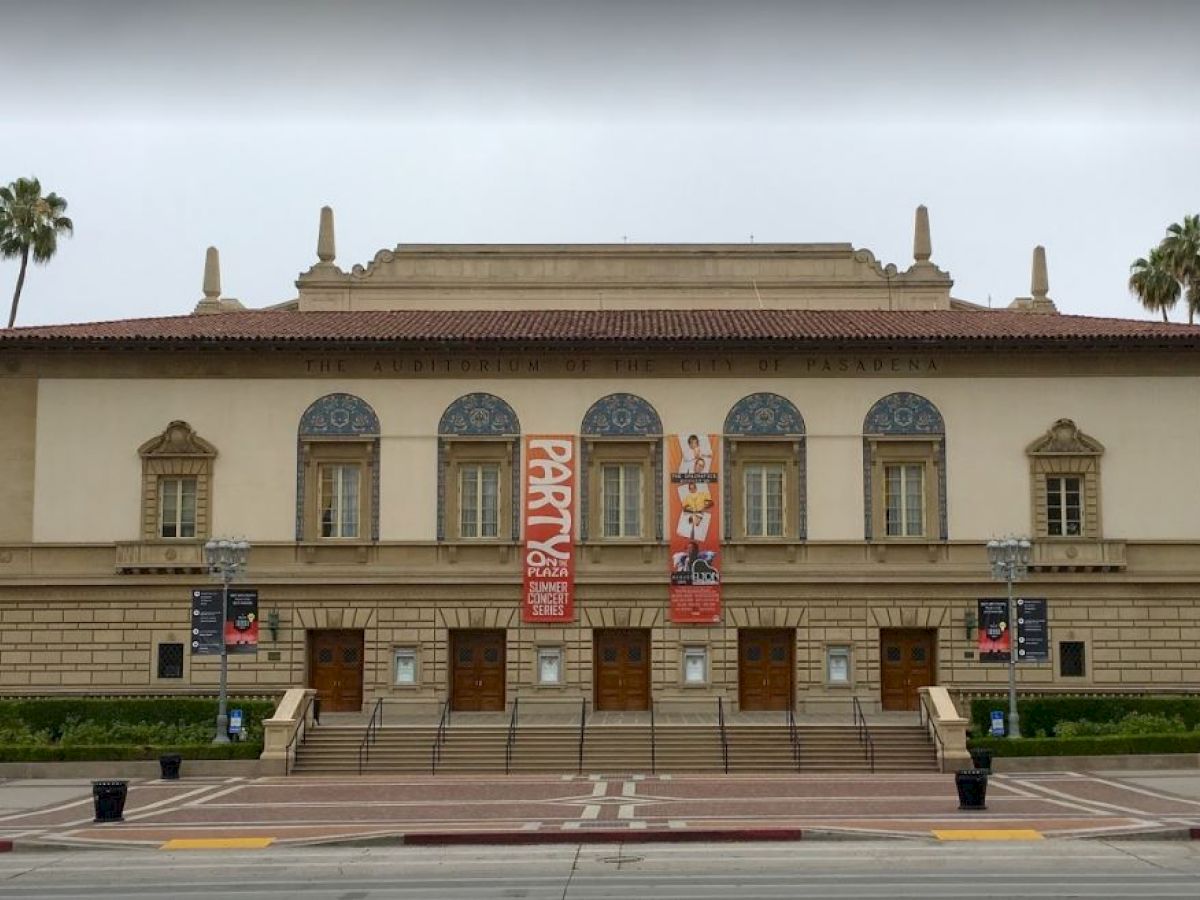 The image shows an ornate building with banners on the front, palm trees, and a wide sidewalk.