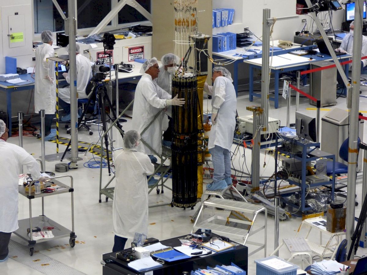 Several people in lab coats and hairnets working on equipment in a lab, surrounded by various tools, electronics, and workstations.