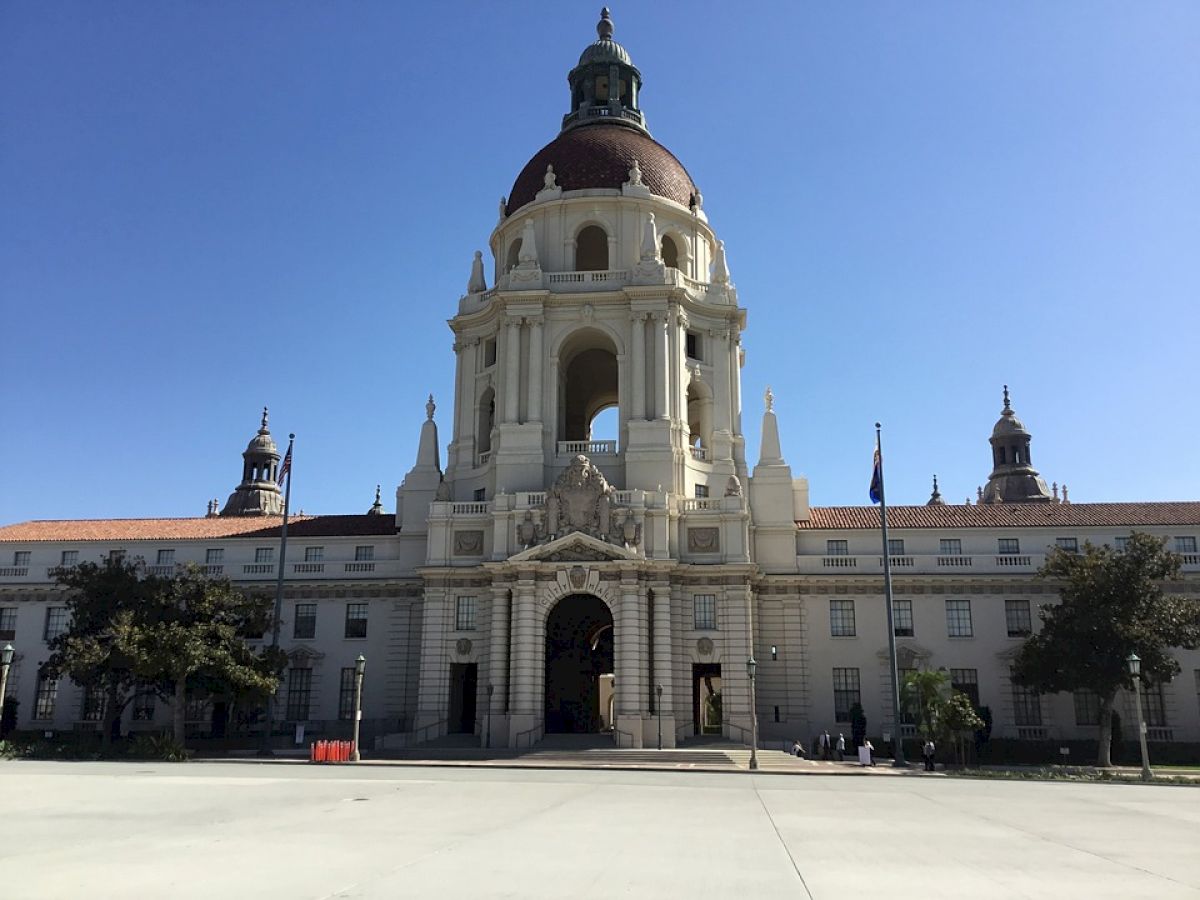 The image shows a large, historic building with a dome and arched entrance, flanked by smaller towers, and trees on either side.