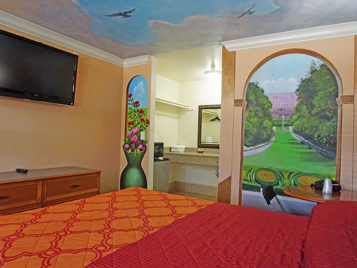 A cozy hotel room with a bed, dresser, wall-mounted TV, colorful wall murals, and a view of a landscaped path through an archway.