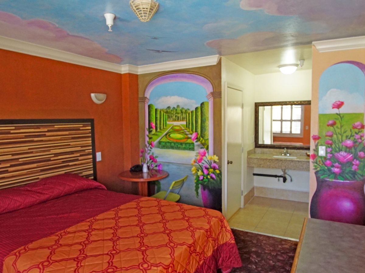 A hotel room contains a bed with red bedding, colorful murals on the walls, a small table, a sink area, and a mounted television.