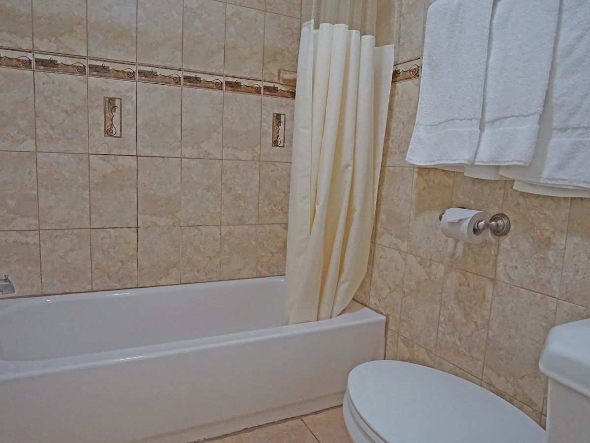 This image shows a bathroom with a tiled bathtub and shower combination, a shower curtain, white towels, a toilet, and a portion of a sink.