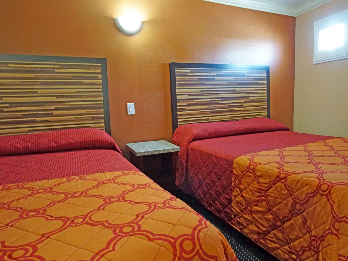 The image shows a hotel room with two beds, a brown and orange color scheme, and a nightstand between the beds under a wall-mounted light.