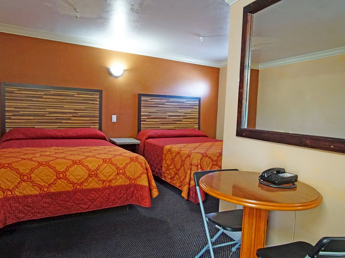 The image shows a hotel room with two beds, a small table with two chairs, a phone, and a large wall mirror.