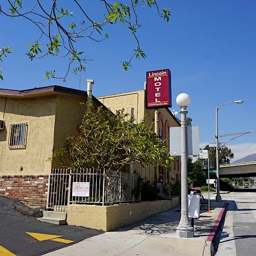 The image shows a motel with a red sign, located on a sunny street corner near a freeway overpass and surrounded by mountains in the background.