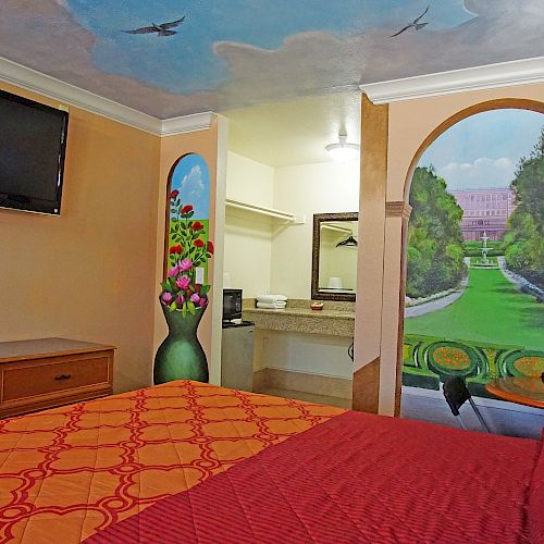 The image shows a hotel room with a wall mural of a garden, a bed with red linens, a wall-mounted TV, and a vanity area with a sink.