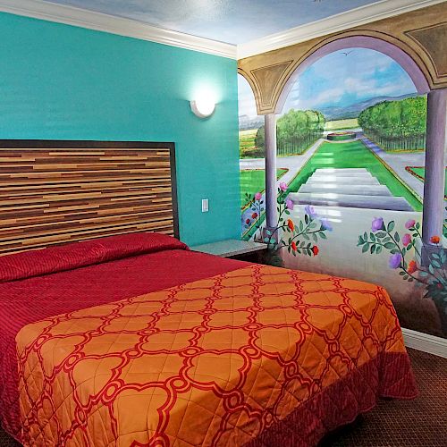 A bedroom with a double bed, bright bedspread, colorful wall mural of a garden, blue walls, light fixtures, and a glimpse of a bathroom.