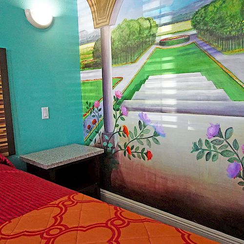 The image shows a small, colorful bedroom with a bed and a vibrant mural of a garden on the wall next to it, creating a scenic atmosphere.