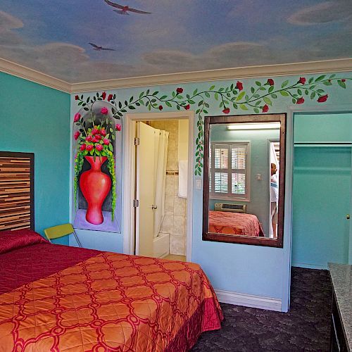 A brightly colored room with a mural above the bed, a large mirror, a door to a bathroom, a TV on the wall, and an orange patterned bedspread.