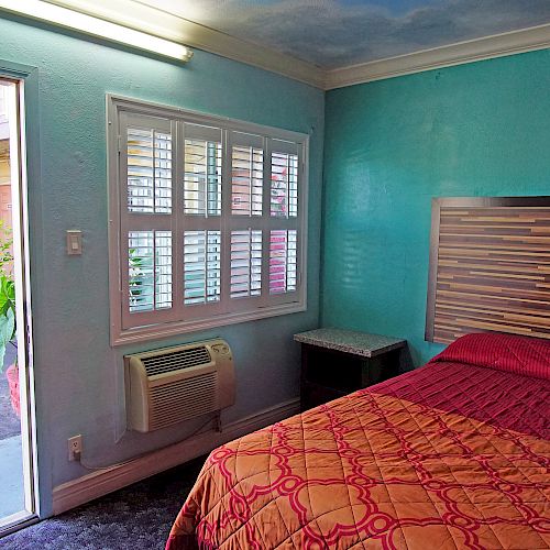 This image shows a motel room with a simple bed, a window with blinds, a small bedside table, and an open door leading to an outdoor corridor.