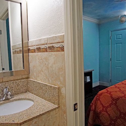 The image shows a small hotel room with a sink and mirror in the bathroom, and an adjacent sleeping area with a bed covered in an orange quilt.