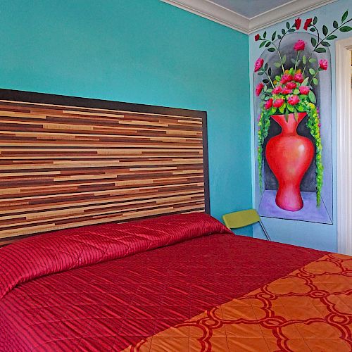 The image shows a brightly colored bedroom with a wooden headboard, red and orange bedding, floral wall art, and a partially open door to a bathroom.