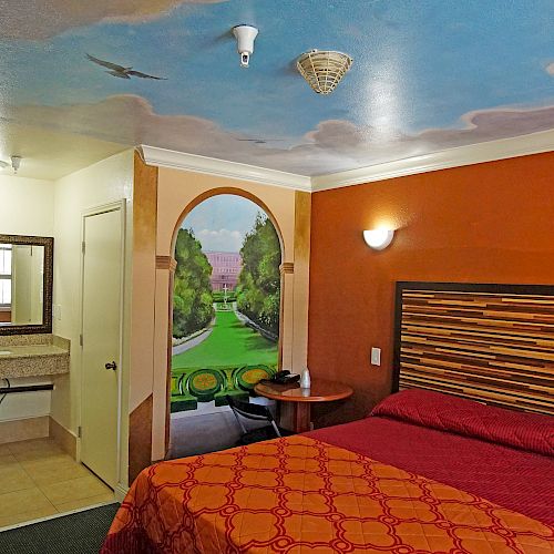 A hotel room with vibrant mural artwork, a red bedspread, a small round table, and a sink area near the bathroom.