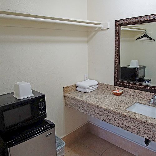 The image shows a hotel room corner with a sink, a framed mirror, a microwave, a mini-fridge, and a shelf with hangers and towels.