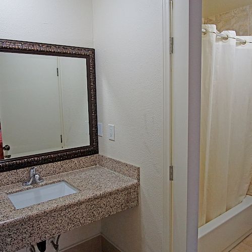 This image shows a bathroom with a granite countertop sink, a large framed mirror, a toilet, and a shower with a beige curtain.