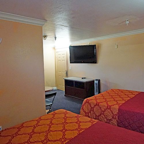 The image shows a hotel room with two beds covered in orange and red quilts. A wall-mounted TV, a small table, chairs, and a fan are visible.