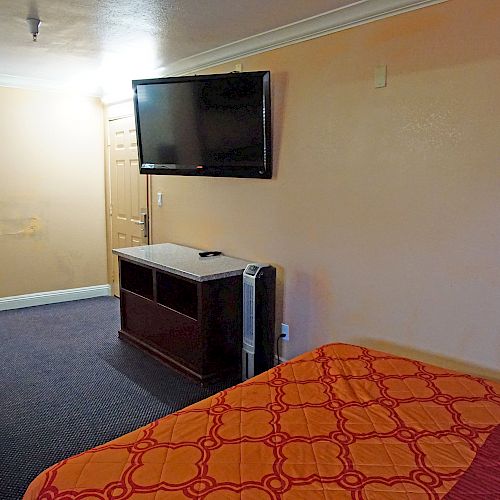 The image shows a small hotel room with a bed, a wall-mounted TV, a table with chairs, an air conditioning unit, and a small window.