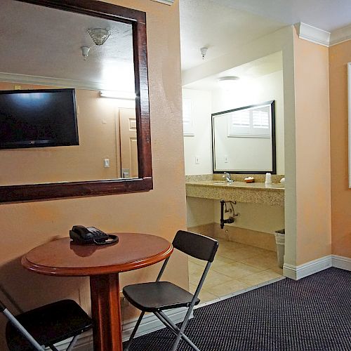 The image shows a small room with a table, two chairs, a wall-mounted TV, a mirror, an air conditioner, and a sink area.