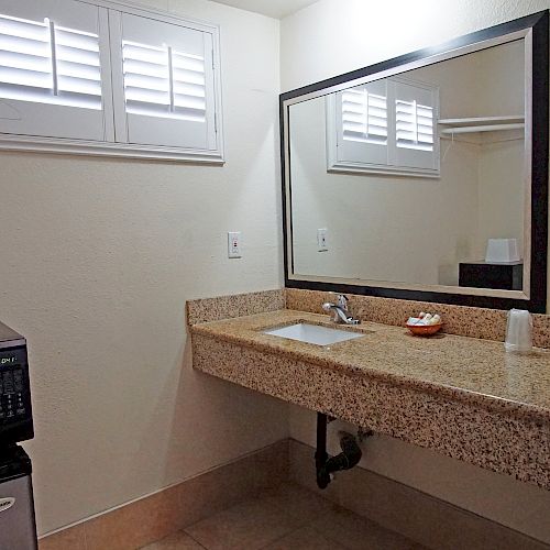 The image shows a bathroom or vanity area with a sink, large mirror, microwave, mini-fridge, towel, and some toiletries on the countertop.