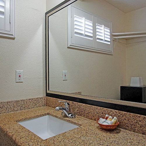 A bathroom sink with a large mirror, granite countertop, soap and lotion basket, a towel, and a reflection of a microwave and a lamp.