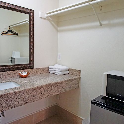 The image shows a room with a sink counter, a mirror, a microwave, a mini-fridge, towels, and a clothes hanger rod on the wall.