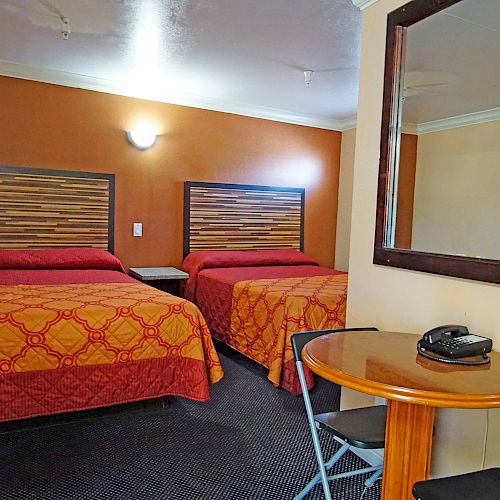 The image shows a hotel room with two double beds, a round table with two chairs, a telephone, and a large mirror on the wall.