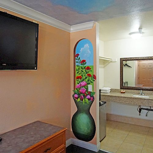 A hotel room features a wall-mounted TV, a dresser, artwork of a vase with flowers, and a bathroom sink with a mirror in the background.