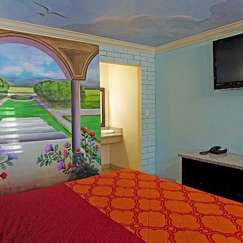 A bedroom features a mural of a garden, a bed with red and orange bedding, a TV on the wall, and a small table with a phone and other items ending the sentence.