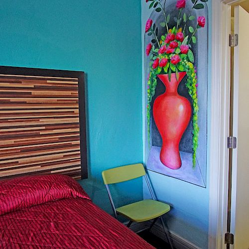 A small bedroom with a striped headboard, red bedding, a yellow chair, a painting of a red vase with flowers, and an open door leading to a bathroom.