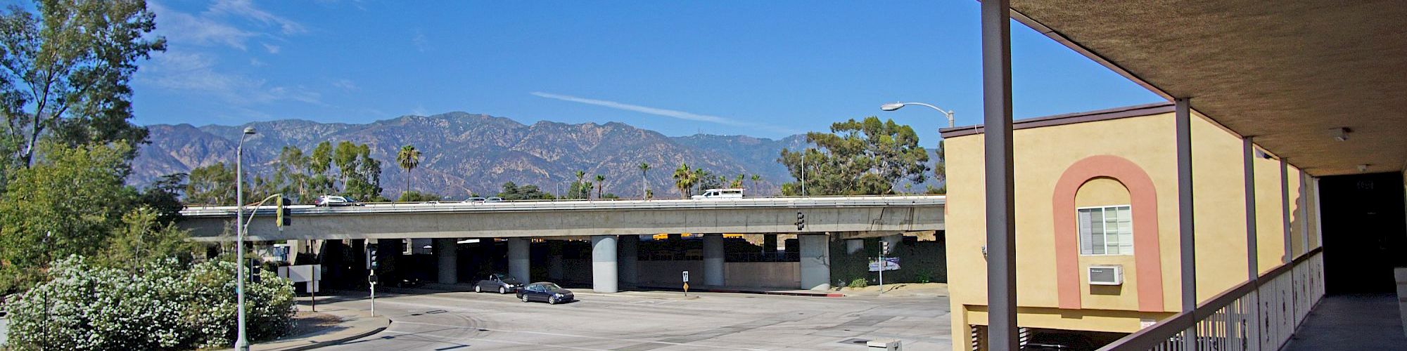 The image shows a view from a balcony overlooking a street with an overpass and greenery against a backdrop of mountains on a clear day.