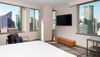 A modern bedroom featuring large windows with a city view, a mounted TV, a chair with cushions, and minimalistic furniture.