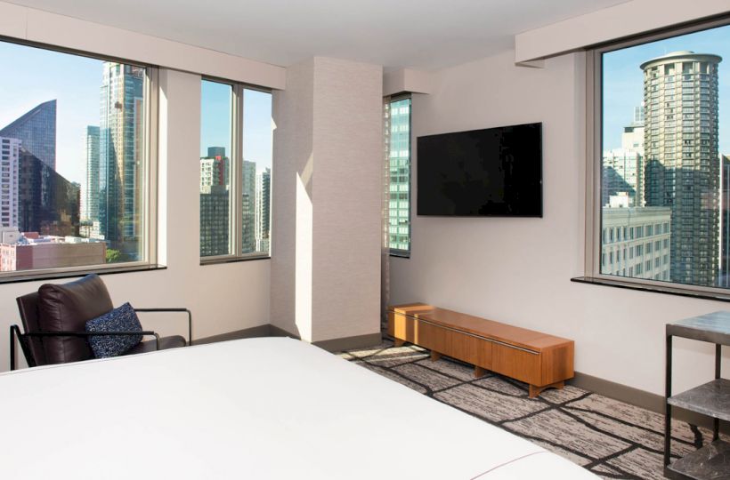A modern bedroom featuring large windows with a city view, a mounted TV, a chair with cushions, and minimalistic furniture.