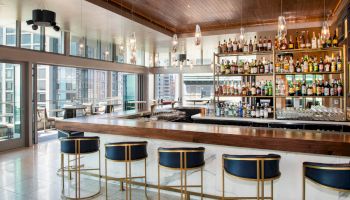 A modern bar with high-back stools, a variety of bottles on shelves, and large windows offering a city view. The atmosphere is chic and inviting.