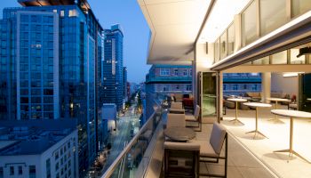 The image shows a modern balcony overlooking a cityscape with tall buildings. The balcony has seating and tables, and it's evening time.
