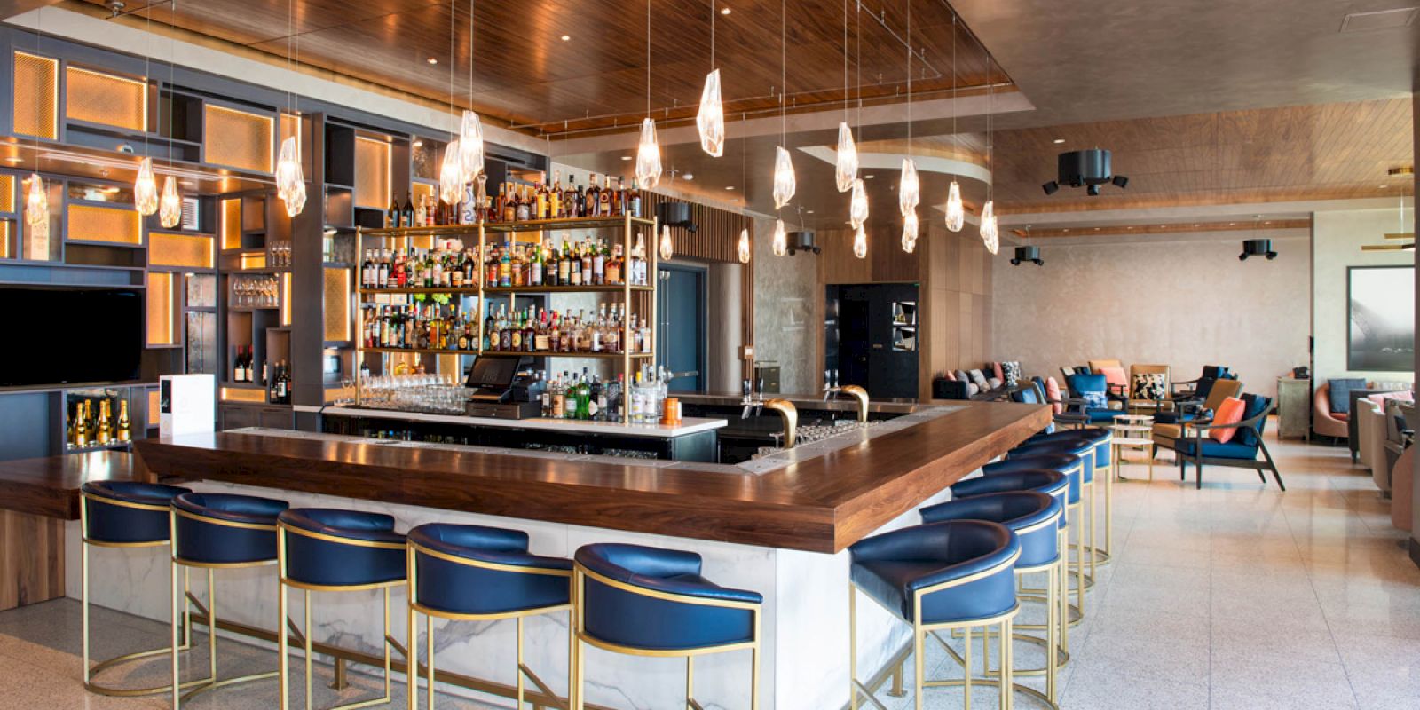 The image shows a modern bar with a central counter, blue bar stools, hanging pendant lights, and a well-stocked shelf of various bottles.