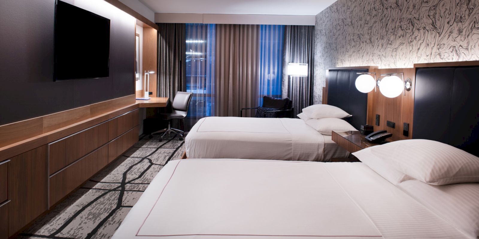 A modern hotel room with two double beds, a flat-screen TV, a work desk, and floor-to-ceiling curtains on the window, with stylish decor.