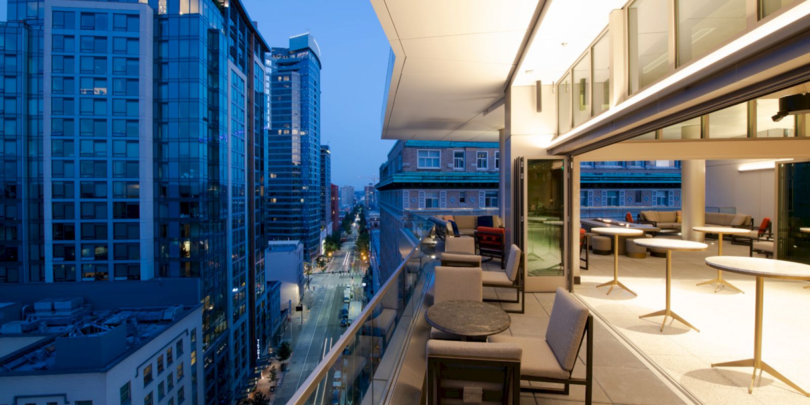 A modern balcony with seating and tables overlooks a city street lined with tall buildings during twilight, showcasing the urban view.