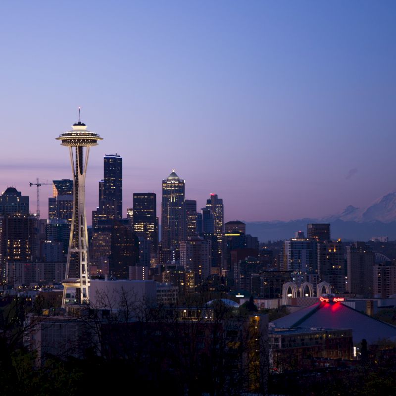 This image shows the Seattle skyline at dusk, featuring the Space Needle and Mount Rainier in the background against a purple and blue sky.