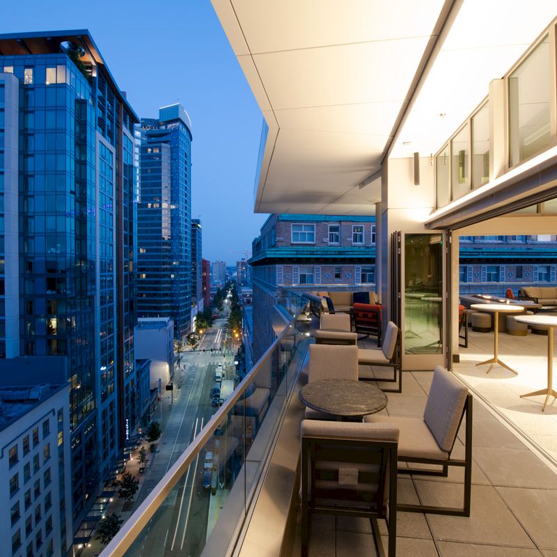 A modern balcony with tables and chairs overlooks a city street lined with tall buildings at dusk, featuring an inviting ambiance.