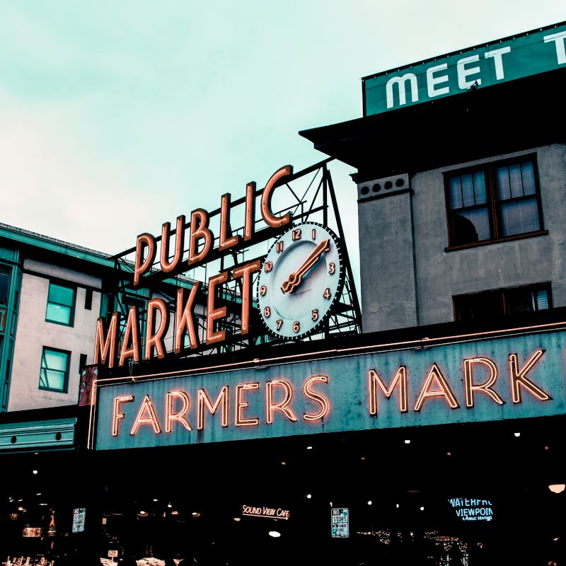 The image shows the Public Market and Farmers Market signs at Pike Place Market in Seattle, with a clock and surrounding buildings.
