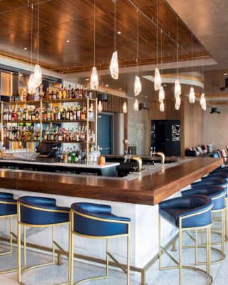 A modern bar with a sleek design, featuring a wooden counter, blue bar stools, hanging pendant lights, and shelves stocked with various bottles.