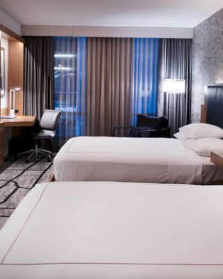 This image shows a modern hotel room with two beds, a large TV on the wall, a desk with a chair, and contemporary decor and lighting.