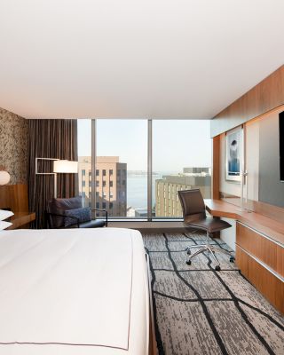 A modern hotel room with a large bed, flat-screen TV, desk, chair, armchair, and window with a city view.