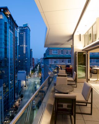 A modern balcony view at dusk overlooking a city street and tall buildings, with seating and tables arranged along the spacious outdoor area.