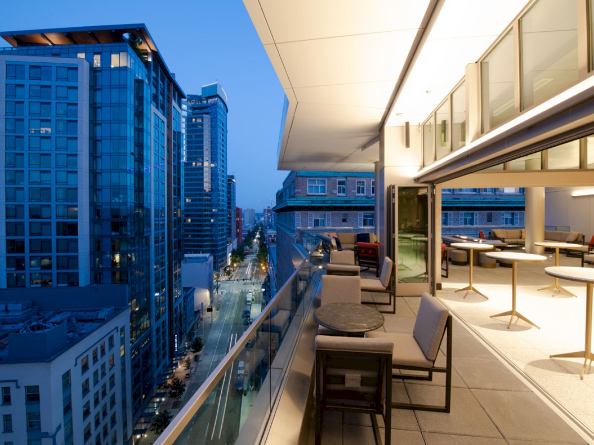 A modern cityscape at dusk, with high-rise buildings and a sleek, furnished balcony overlooking a street, featuring tables, chairs, and ambient lighting.