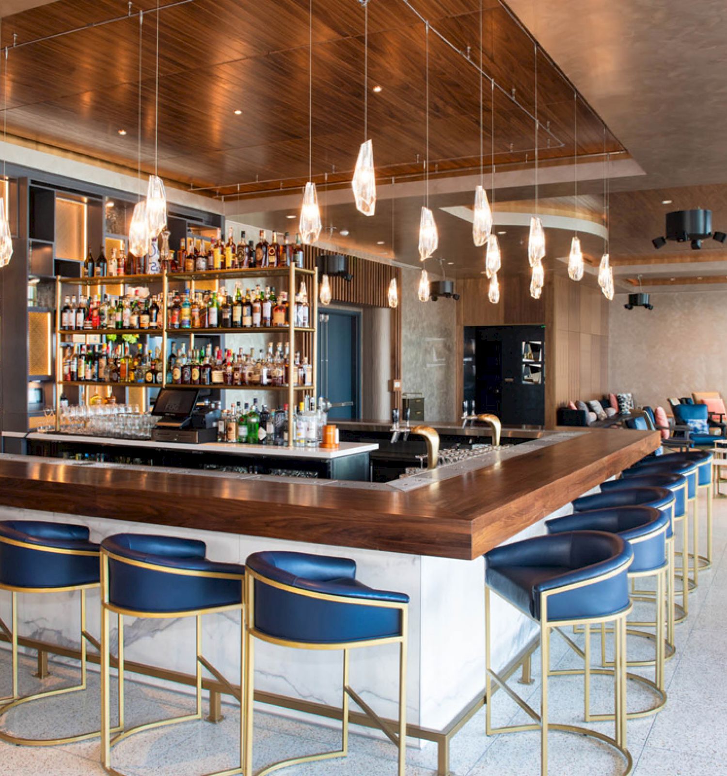 The image shows a modern bar with a wooden counter, blue cushioned bar stools, hanging pendant lights, and a variety of bottles arranged on shelves.
